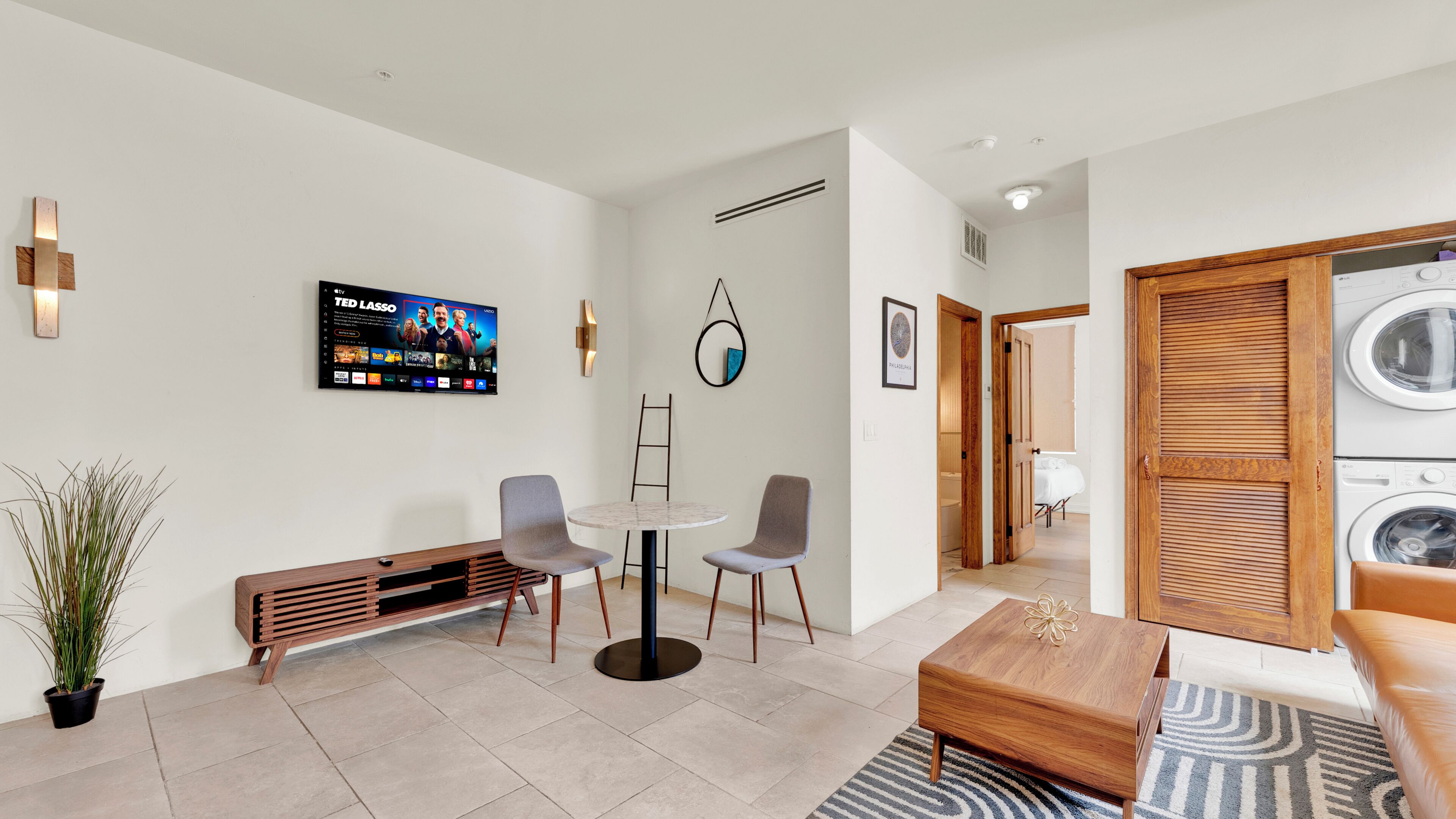 Une chambre d’hôtel moderne équipée d’un téléviseur à écran plat, d’un banc en bois, de deux chaises, d’une table basse, d’une plante en pot et d’un espace dédié au linge.