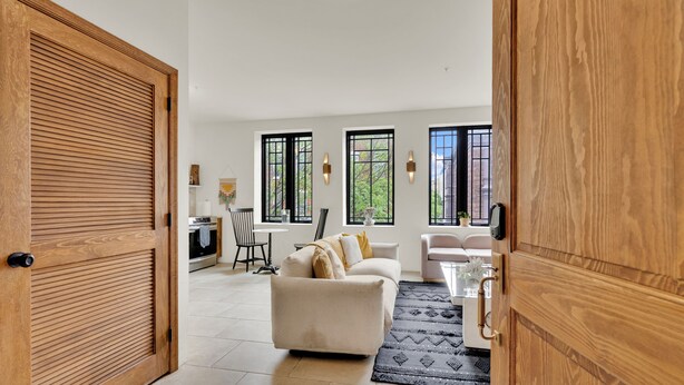 Uma porta de madeira aberta dá acesso a uma sala de estar com sofá, mesa de jantar e área de cozinha visíveis pelas janelas.