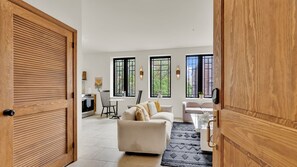 Puerta de madera abierta que da a una sala de estar con sofá, una mesa de comedor y una cocina visible a través de las ventanas.