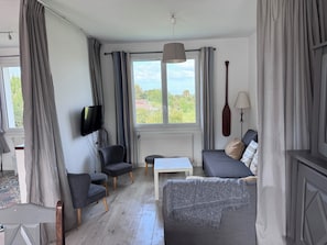 A modern living room with a sofa, armchair, TV, and a view of greenery outside. - Villa Jo - Family Home in Grand-Village (Le Grand-Village-Plage)