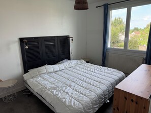 A bedroom with a bed, a small round table, a wooden desk, a radiator, and a window with a view of greenery. - Villa Jo - Family Home in Grand-Village (Le Grand-Village-Plage)