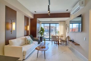 A modern living room with a sofa, dining table, chairs, and a large window with a city view. - ®{2BR | Urban~Oasis} DowntownSD +Pool+Gym+Balcony (Santo Domingo)
