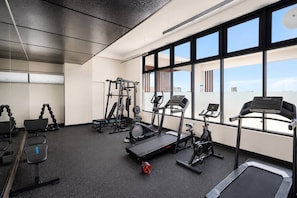 Une salle de sport équipée de divers appareils d’exercice, dotée de grandes fenêtres et offrant une vue sur le paysage urbain.