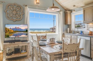 A dining area with a table set for four, a television mounted on a wooden stand, and a view of the ocean through a large window.