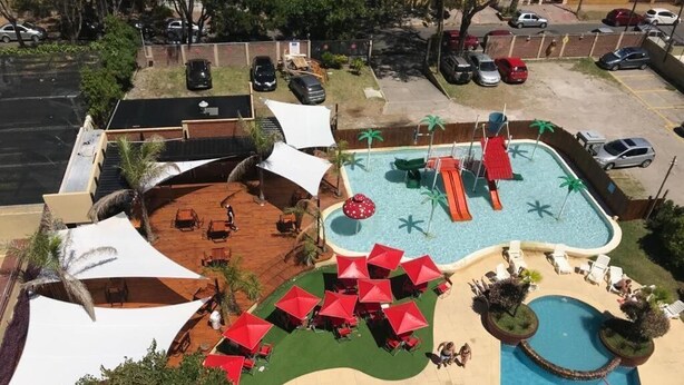 Un área de piscina con múltiples sillones de descanso y sombrillas, un tobogán pequeño y una piscina separada.