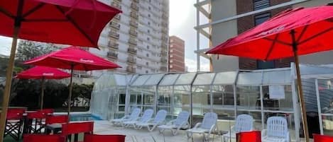 A rooftop pool area with red umbrellas, lounge chairs, and a glass-enclosed structure.