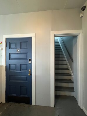 Una puerta azul con teclado electrónico, una escalera con barandilla y una pared blanca con detector de humo.