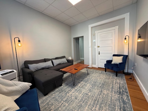 Un salon comprenant un canapé, un pouf, une table basse et un fauteuil bleu. Un téléviseur à écran plat est fixé au mur, et une porte donne accès à une autre pièce.