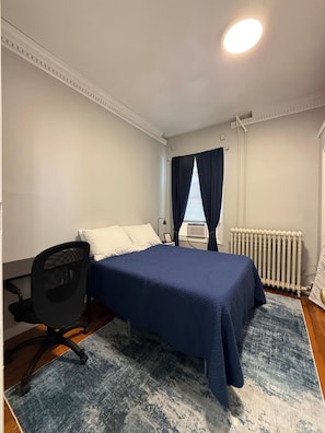 Un dormitorio con cama, escritorio, silla, radiador y ventana con cortinas.