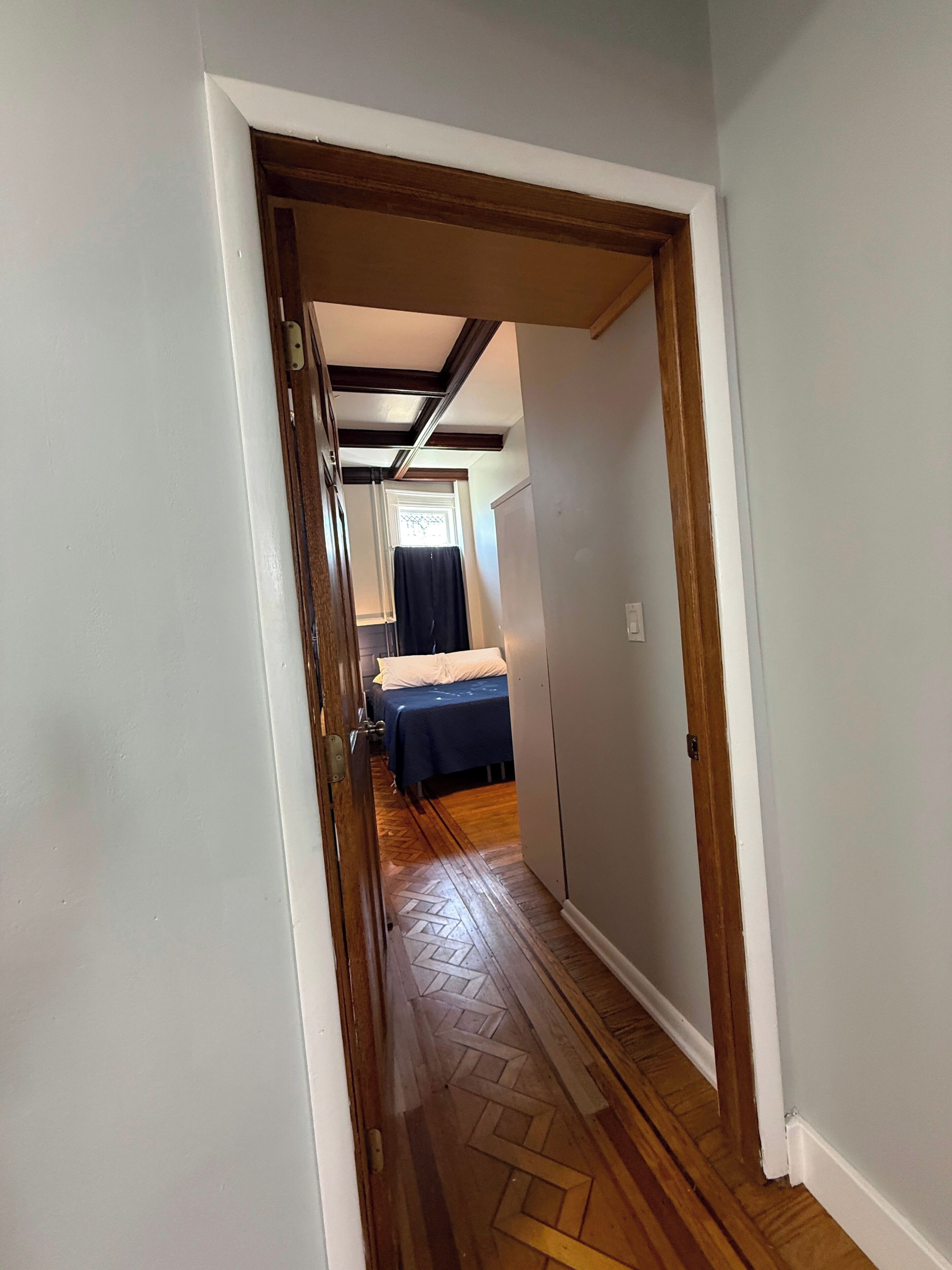 A narrow hallway with wooden flooring leading to a room with a bed and a window.