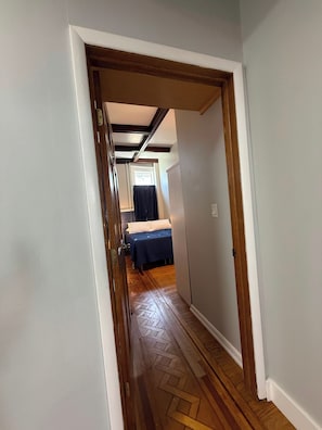 A narrow hallway with wooden flooring leading to a room with a bed and a window.