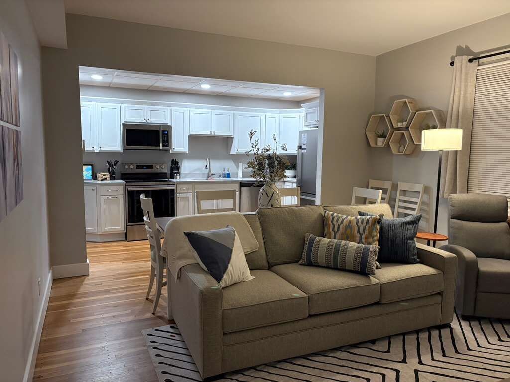A living room with a sofa, a coffee table, and a dining area with chairs. There is a kitchen in the background with white cabinets and appliances.