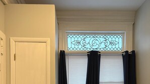 A room with a radiator, a window with curtains, a fan, and a bed with a patterned bedspread. - DM-A Historic 2BR Apartment (DuBois)