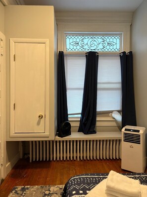 Una habitación con radiador, ventana con cortinas, ventilador y cama con colcha estampada.