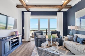 A modern living room with a large window, a sofa, a chair, a coffee table, and a TV. - Oceanfront Resort Retreat | Dual Homes, Luxe Pool & Endless Gathering Space (Emerald Isle)