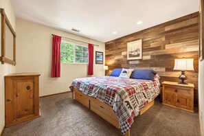 Une chambre à coucher avec une tête de lit en bois, un lit recouvert d’une couverture à carreaux, deux tables de chevet avec des lampes, une fenêtre avec des rideaux rouges et une armoire en bois.