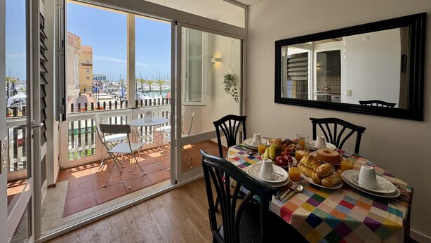 Spiseområde med et bord dækket til morgenmad, en balkon med udsigt over lystbådehavnen og et stort spejl på væggen.