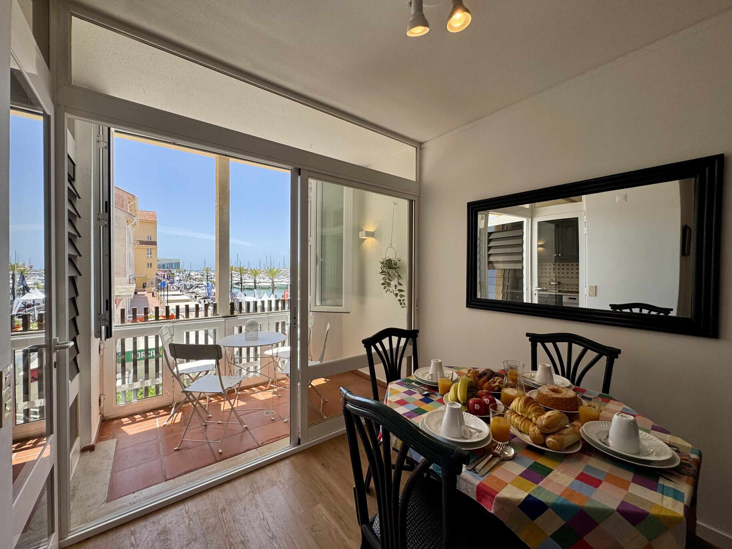 Un comedor con una mesa preparada para el desayuno, un balcón con vistas al puerto deportivo y un espejo grande en la pared.