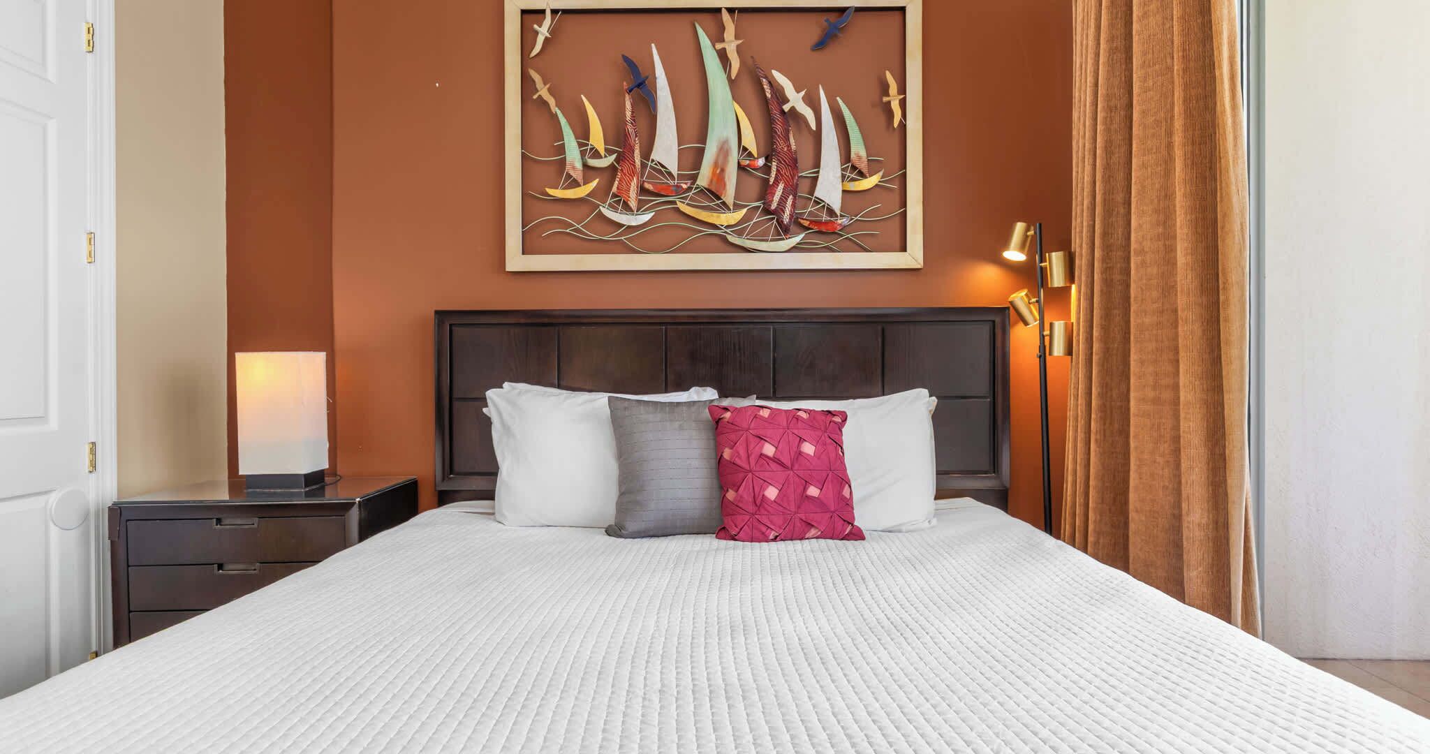 A hotel room with a large bed, a nightstand, a lamp, a painting of sailboats, and a curtain.