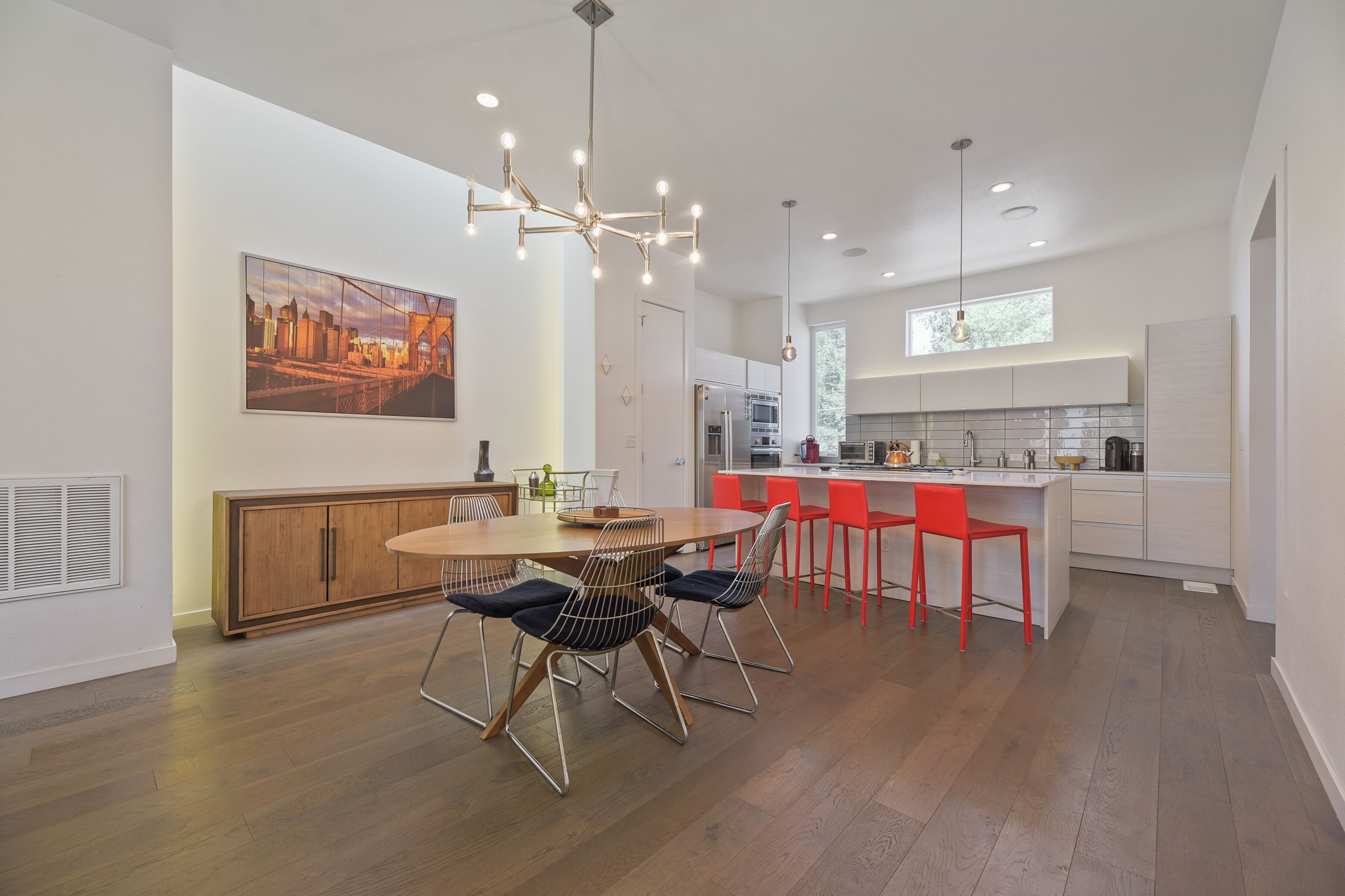 A modern kitchen with a dining area, wooden cabinets, a large painting, and a chandelier.