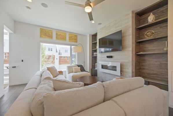 A modern living room with a large sectional sofa, a flat-screen TV mounted on a wooden wall, and a ceiling fan. - Sloan's Lake Luxury | Private Rooftop | 30-Day Min (Denver)