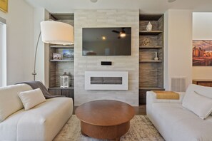 A modern living room with a flat-screen TV mounted on a brick wall, a round wooden coffee table, and two white sofas. - Sloan's Lake Luxury | Private Rooftop | 30-Day Min (Denver)