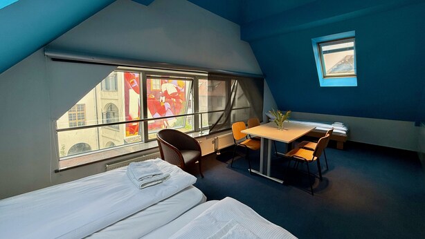A room with two beds, a table with chairs, a small window, and a view of buildings outside. - Mini-Teater Suites - Apartment 6 - sleeps 5 guests  in 2 bedrooms (Ljubljana)