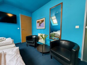 Ein Hotelzimmer mit blauer Wand, einem Bett mit weißen Leinen, zwei schwarzen Ledersesseln, einem kleinen runden Tisch mit einer Vase, einem gerahmten Bild und einem großen Spiegel.