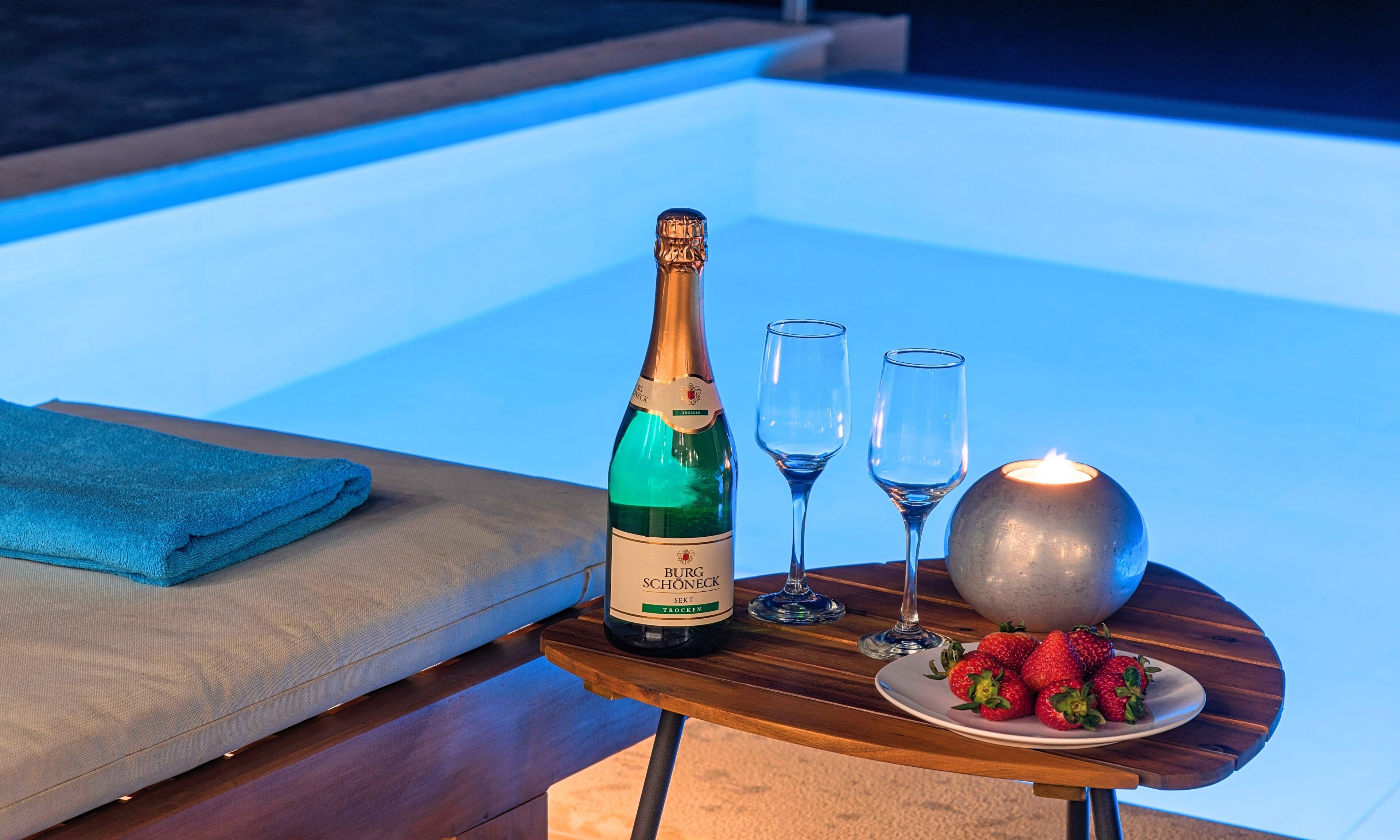 En flaske Burg Schöneck sekt trocken, to glas, et stearinlys og jordbær på et bord ved en pool.