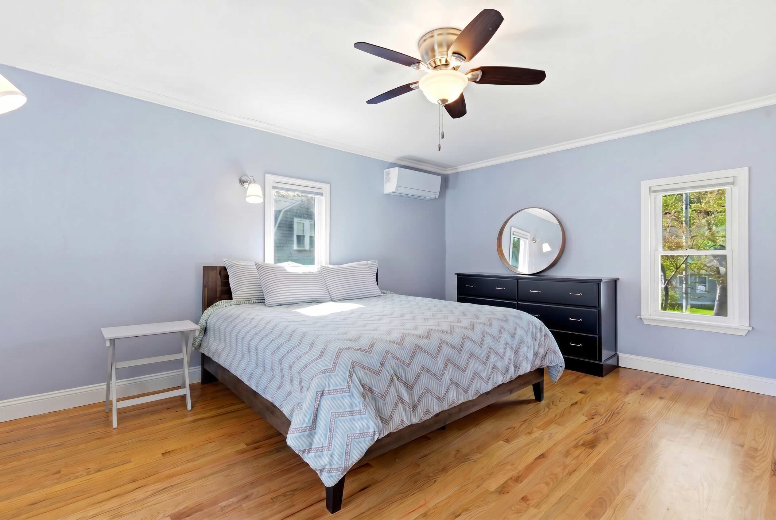 A bedroom with a bed, a ceiling fan, a bedside table, a dresser, a mirror, and a window.