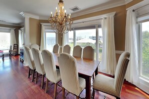A dining area with a large wooden table and six upholstered chairs, a chandelier, and large windows with curtains. - Tybee Nights | Great Views, Steps From Sand, Elevator (Tybee Island)