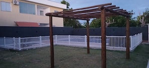 A wooden play structure in a fenced backyard with a building in the background. - Residencial Praia dos Corais (Porto Seguro)