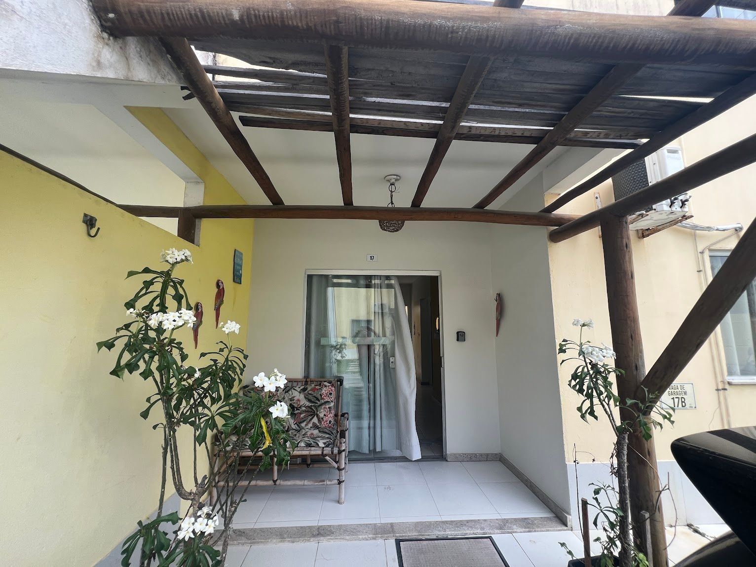 Área externa coberta com um banco, porta de entrada e um vaso de planta.