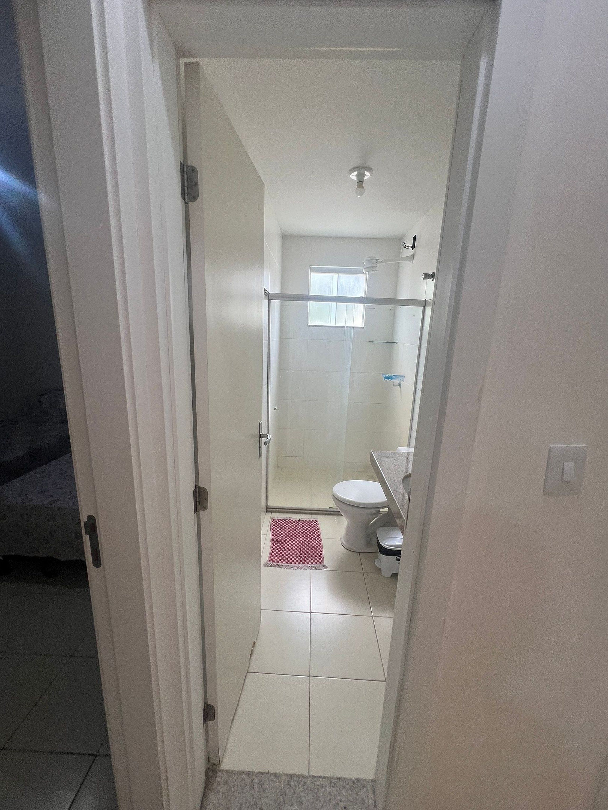 Un couloir étroit mène à une salle de bain équipée d’une douche, de toilettes et d’une petite fenêtre.
