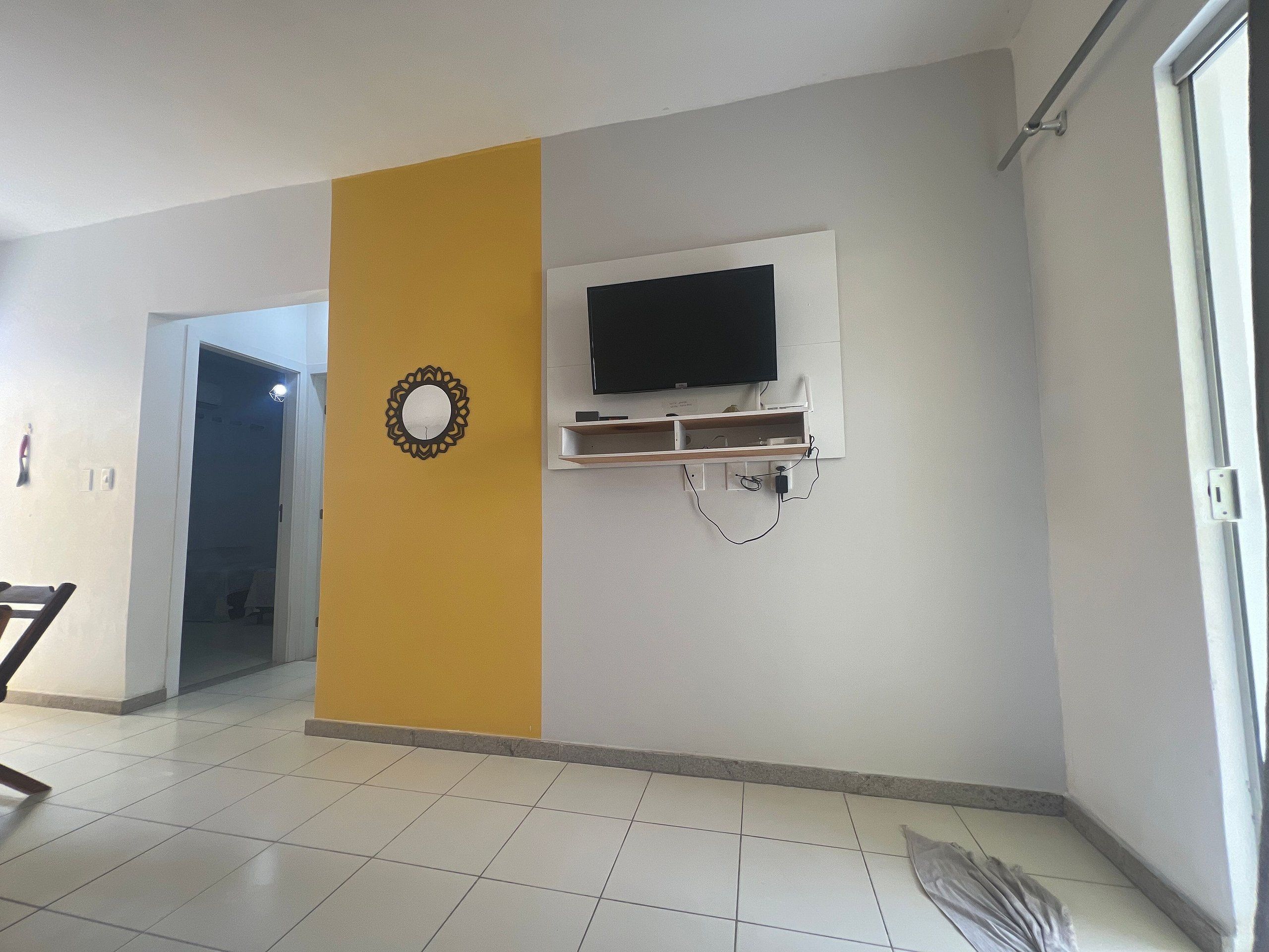 Quarto com televisão na parede, porta para outro cômodo e piso ladrilhado.