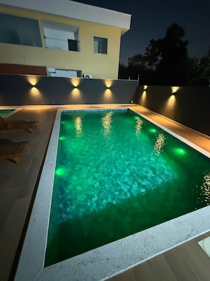 A rectangular swimming pool with clear water, surrounded by a light-colored deck and a modern building in the background. - Residencial Praia dos Corais (Porto Seguro)