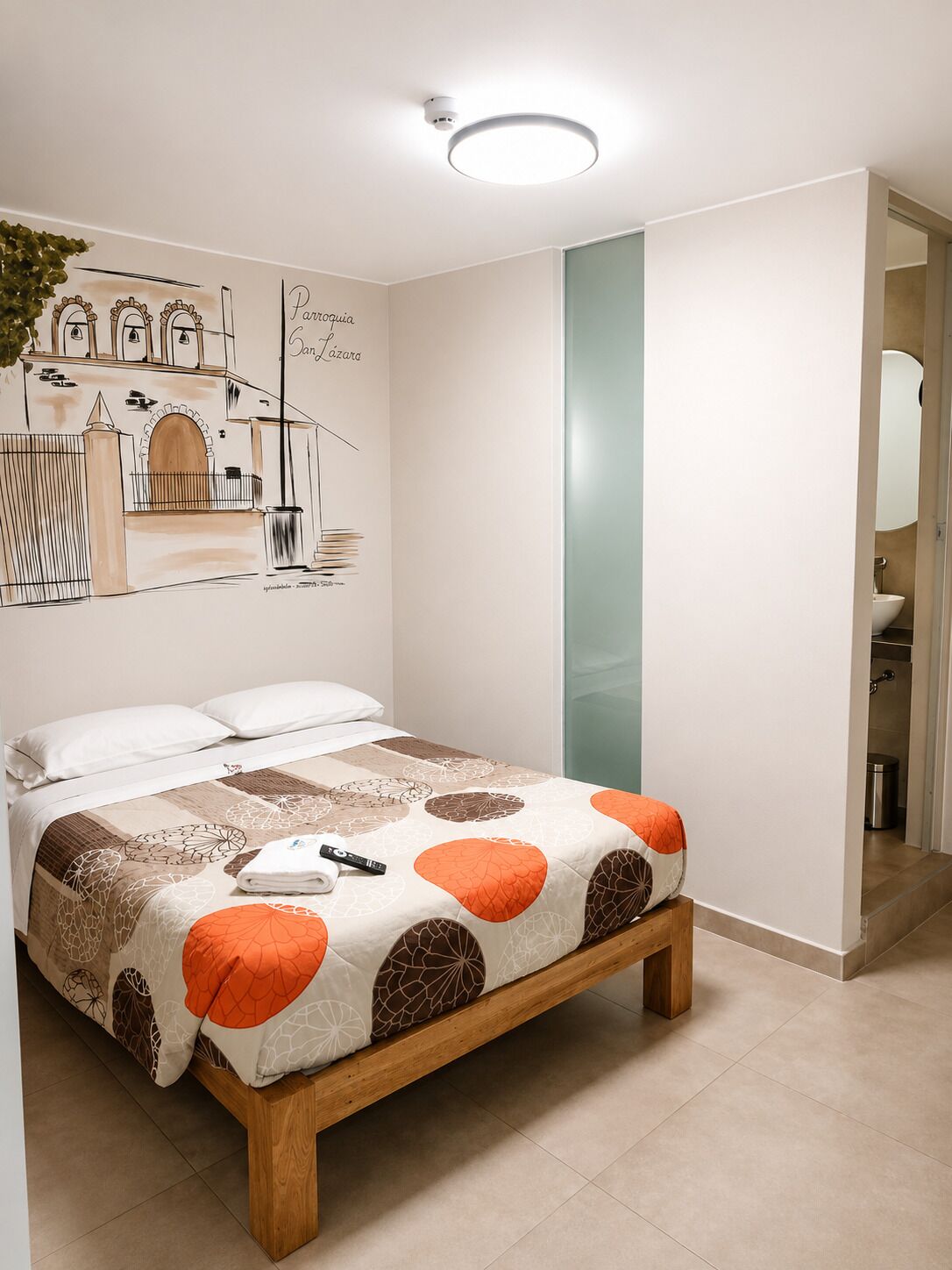 Ein Schlafzimmer mit Bett, Wandgemälde, einem durchgehenden Türrahmen einsehbaren Badezimmer und einer Deckenleuchte.
