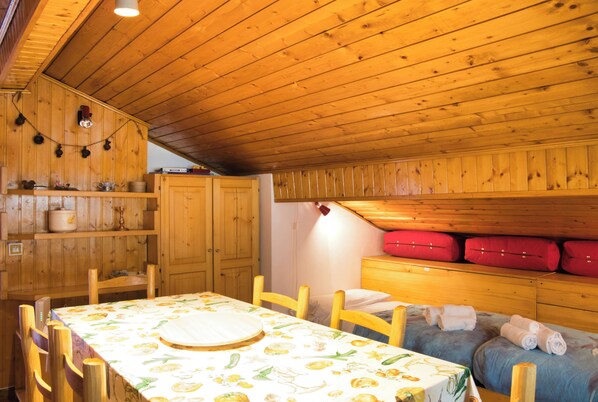 A wooden interior with a dining table, chairs, a small kitchen area, and a sleeping area with pillows and a red suitcase.