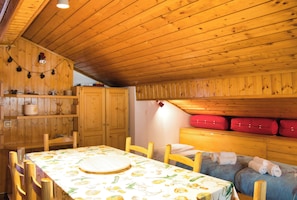 A wooden interior with a dining table, chairs, a small kitchen area, and a sleeping area with pillows and a red suitcase.