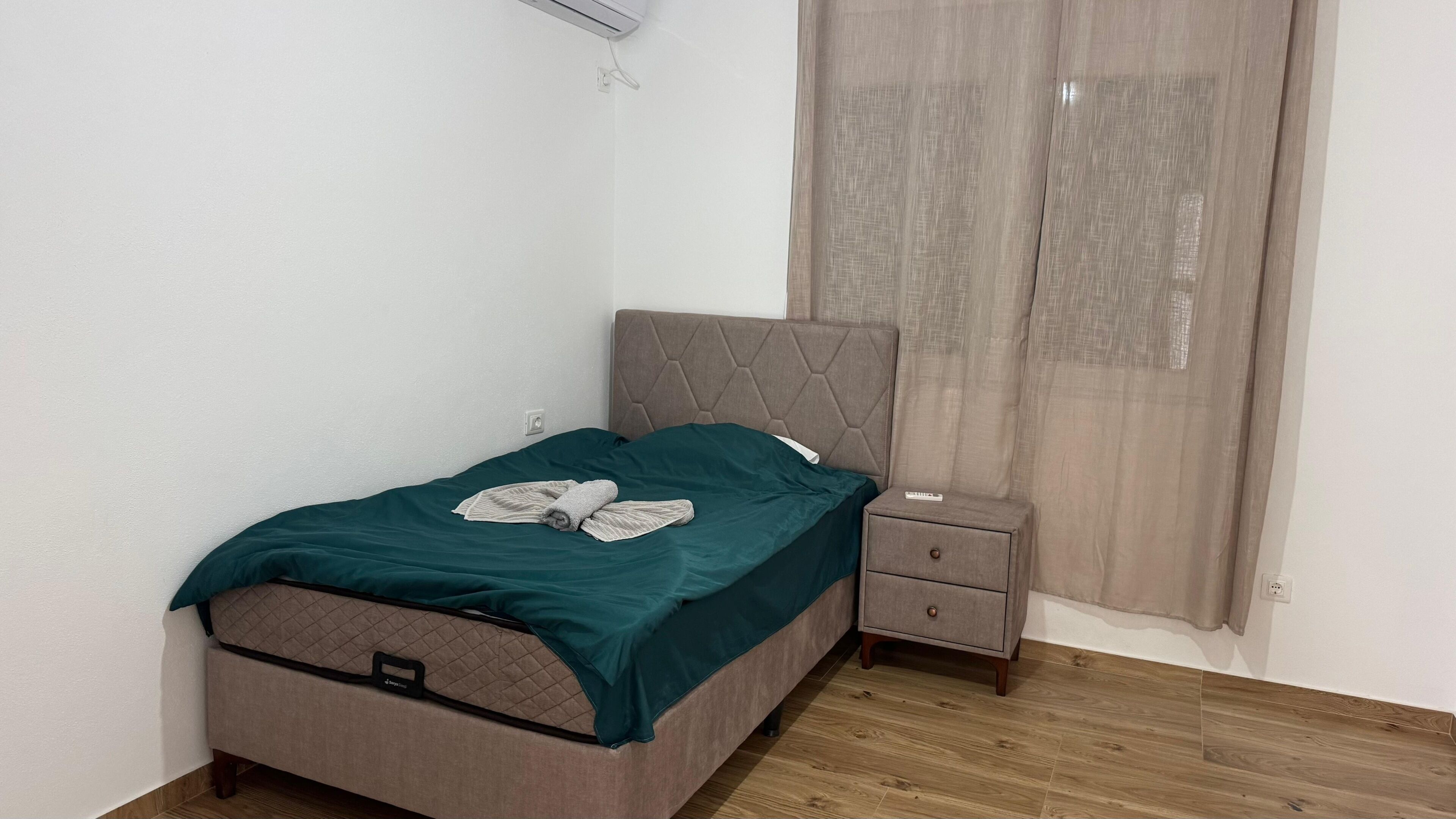 A single bed with a headboard, a bedside table, a small drawer, and a window with curtains.