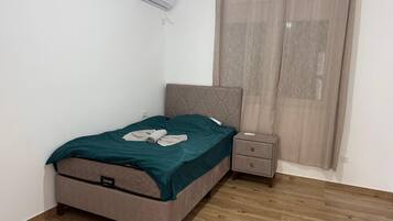 A single bed with a headboard, a bedside table, a small drawer, and a window with curtains.
