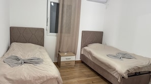 Ein Doppelzimmer mit zwei Betten, einem kleinen Nachttisch, einem Fenster mit Vorhängen und einer Klimaanlage.