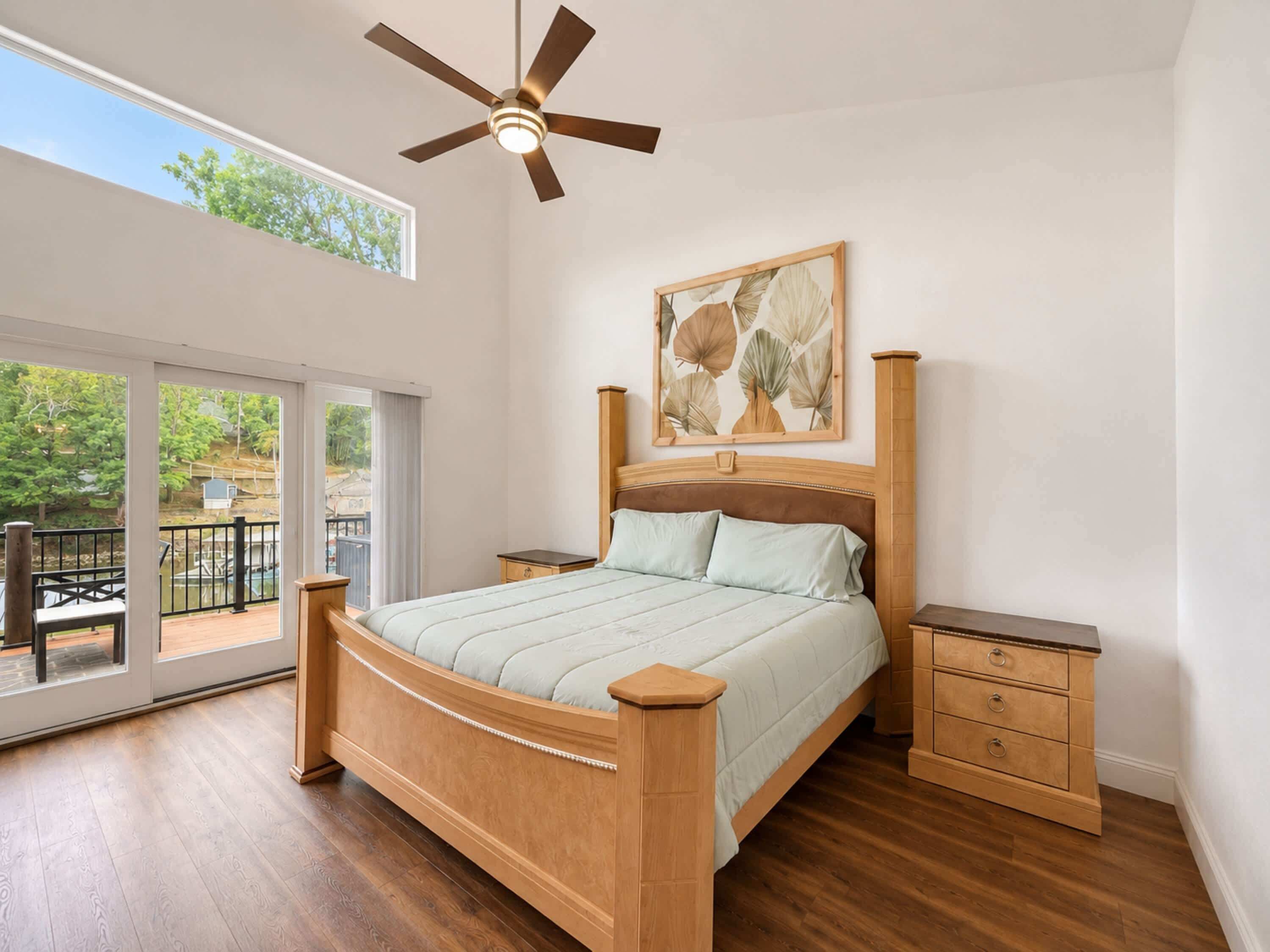 Un dormitorio con una cama grande, un ventilador de techo, una mesita de noche de madera, un aparador de madera y un ventanal corredero que da a un espacio al aire libre.