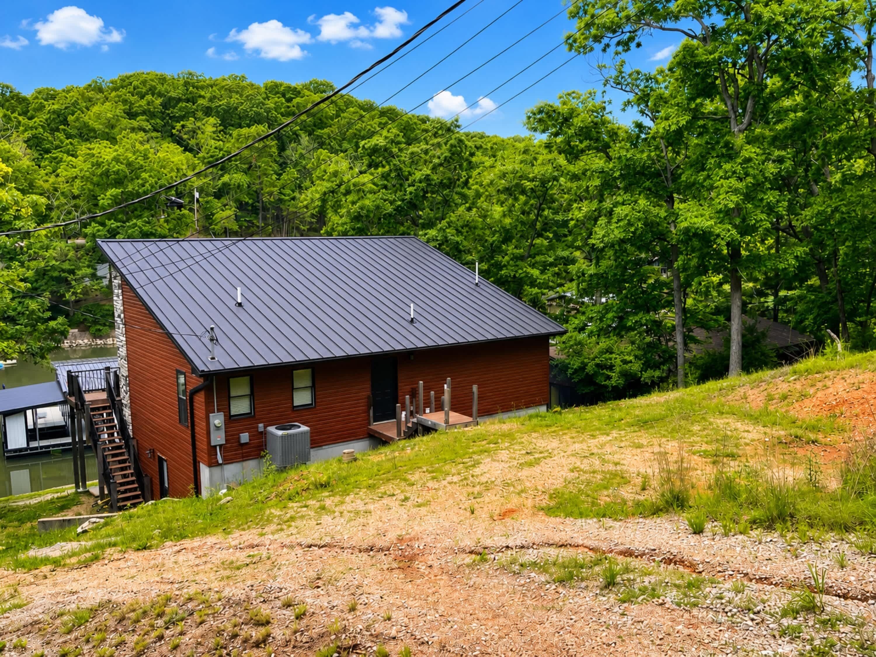 A small, single-story cabin with a metal roof, situated on a grassy hill with a view of a lake and dense trees in the background.