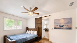 Un dormitorio con cama, piso de madera, ventilador de techo, ventana y puerta que da a otra habitación.