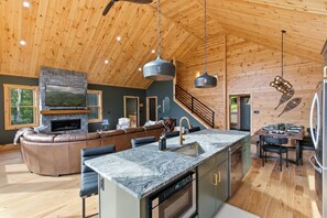 A spacious living area with a stone fireplace, a large leather sofa, and a kitchen island with a sink. - Luxury Chalet w/ Hot Tub, Game Rooms & Mtn Views (Bethel)
