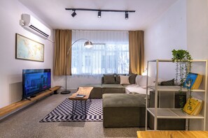 A modern living room with a flat-screen TV, a sofa, a coffee table, a bookshelf, and a large window with curtains.