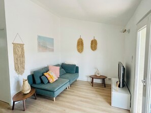 A modern living room with a teal sofa, a small wooden side table, a decorative macrame wall hanging, a round wooden side table, and a wall-mounted television. - Magnificent, peaceful ground floor villa with unobstructed view. Ideal for families (Sainte-Maxime)