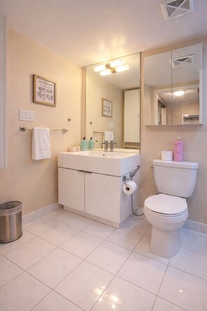 Ein Badezimmer mit weißem Waschbecken, Toilette und Spiegel. An der Wand hängen zwei Bilder, ein Handtuchhalter und ein Mülleimer.