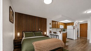Een compacte slaapkamer met een bed, nachtkastjes, een wand van houten latten, een kitchenette en een deur naar een andere kamer.